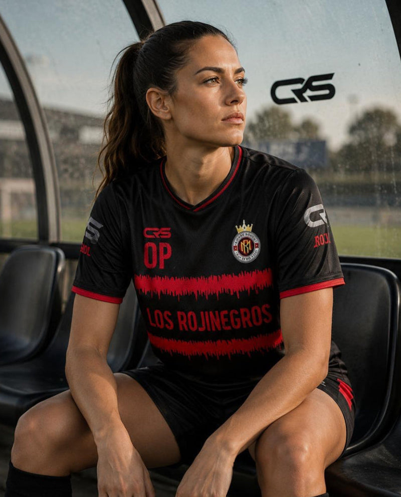 A woman sits on a bench inside a sports stadium shelter wearing a black Los Rojinegros soccer jersey with red accents and black socks. The jersey features the CRS brand logo and the team crest.