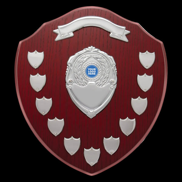 A shield-shaped wooden plaque with a dark red wood grain finish and multiple silver metal badge holders, including one central crest with a blue placeholder logo area and a banner at the top, shown on a plain white background.